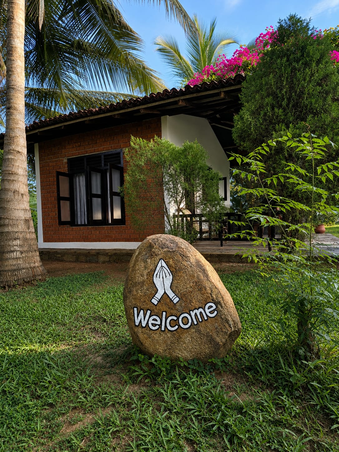 Welcome stone with praying hands in front of a brick bungalow and coconut palms
